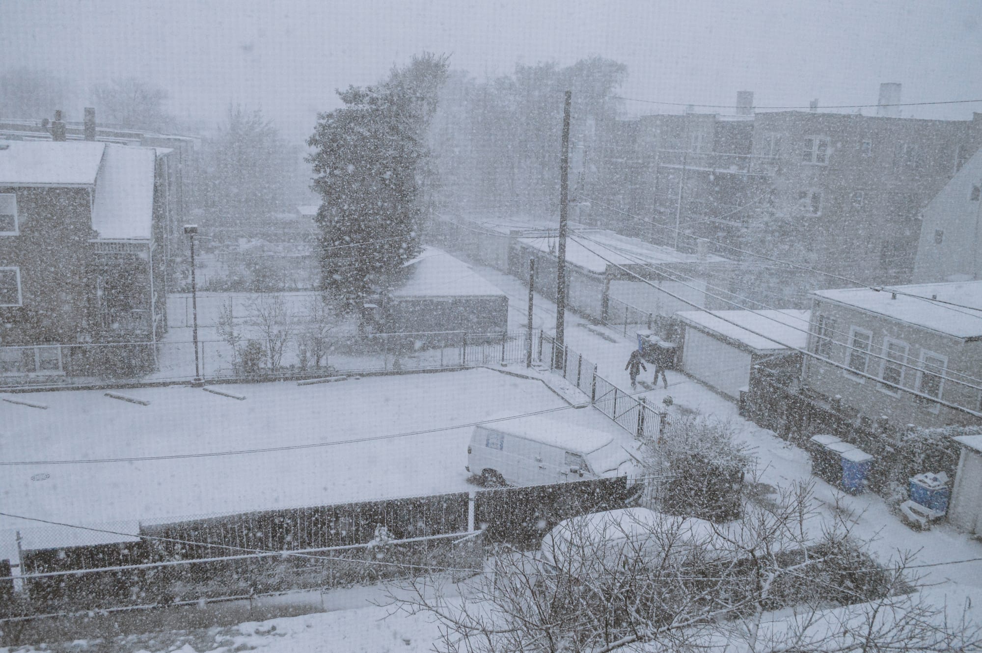 A snow-covered parking lot and alley in a Chicago neighborhood. There are two people walking through the snow from a distance.